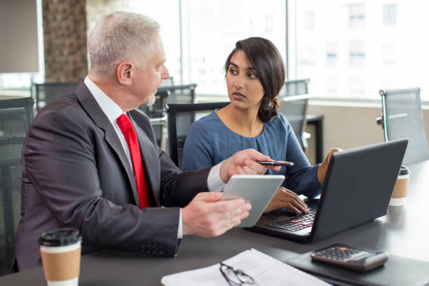 Two business colleagues working together in meeting space. Mentor instructing newcomer. Young Indian woman with laptop and mid adult man with tablet discussing new project. Teamwork or training concept